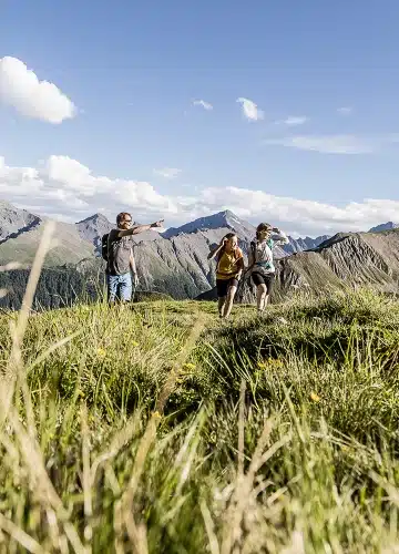 Drei Personen wandern in einer grasbewachsenen Berglandschaft unter einem blauen Himmel mit vereinzelten Wolken.