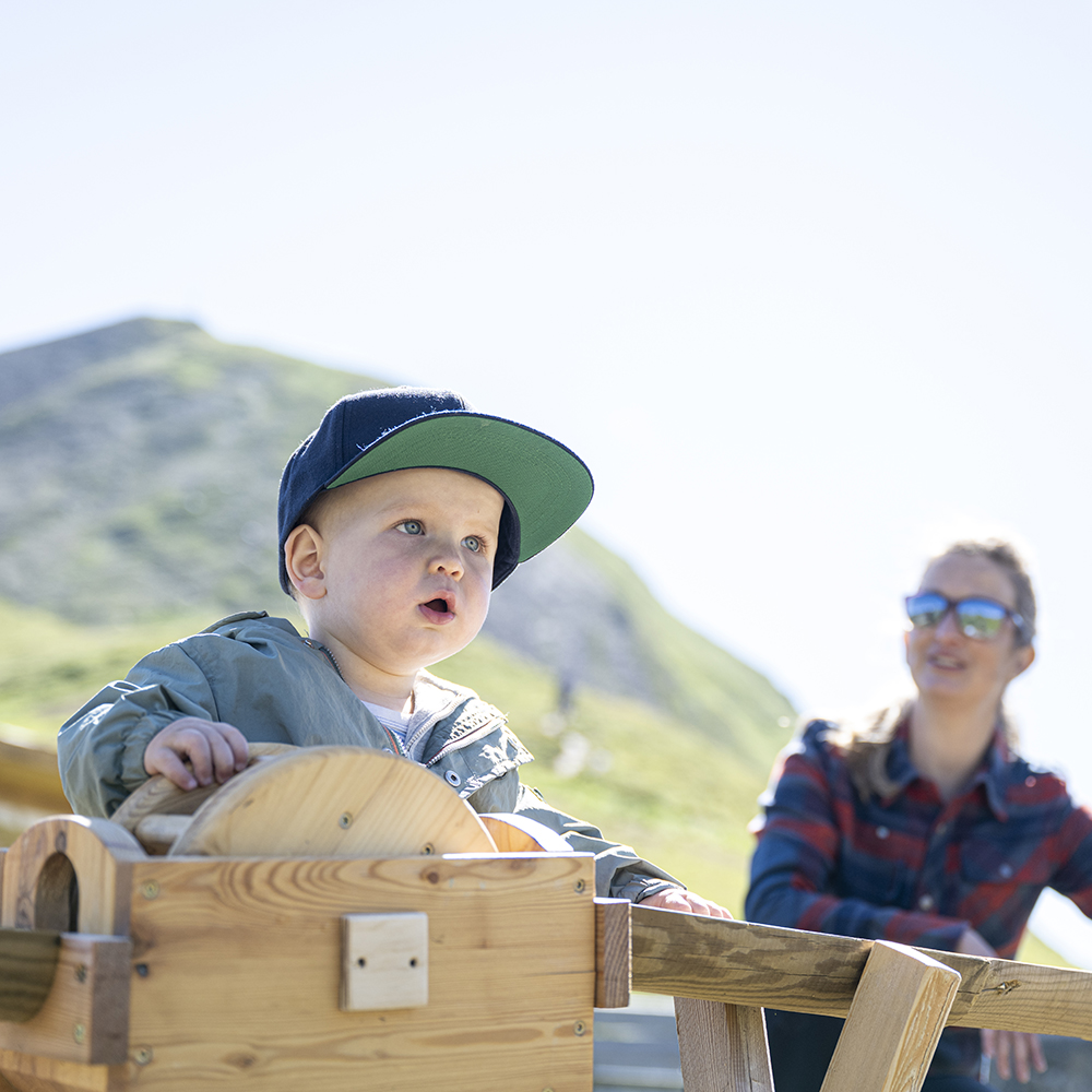 Kleinkind mit Mütze beim Spielen im Freien in Samnaun mit einer Frau im Hintergrund an einem sonnigen Tag.