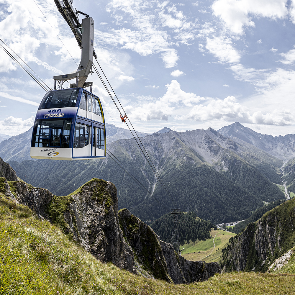 Eine Seilbahn fährt über alpine Berge mit grünen Tälern und fernen Gipfeln unter uns.