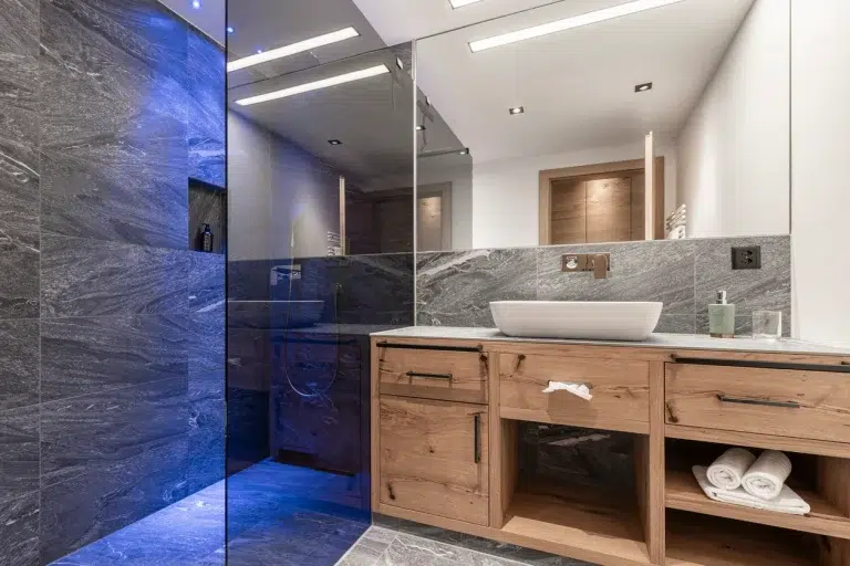 Modernes Badezimmer im Ferienhaus Samnaun mit grauen Steinwänden, Holzwaschtisch und blauer Beleuchtung.
