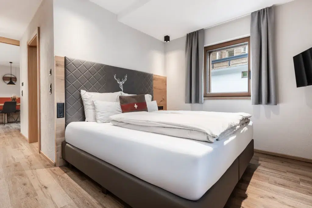 Modernes Schlafzimmer im Ferienhaus Samnaun mit Doppelbett, grauem Kopfteil und wandmontiertem TV.