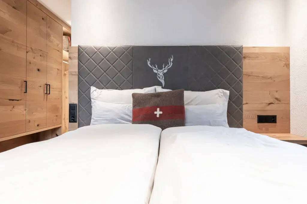 Modernes Bett mit weissem Bettzeug, Hirschkopfteil, Kissen mit Schweizer Flagge im Ferienhaus Samnaun.