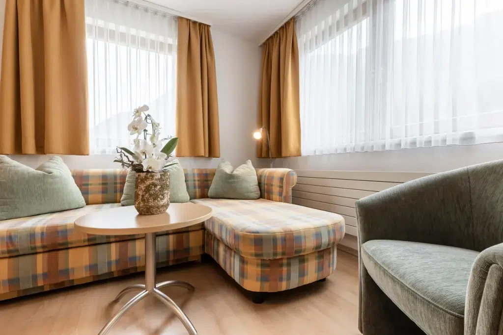 Gemütliches Wohnzimmer in der Ferienwohnung Samnaun mit kariertem Sofa, rundem Tisch und goldenen Vorhängen.