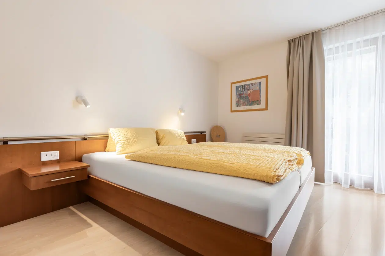 Minimalistisches Schlafzimmer in der Ferienwohnung Samnaun mit gelbem Bettzeug, Holzmöbeln und Sonnenlicht.