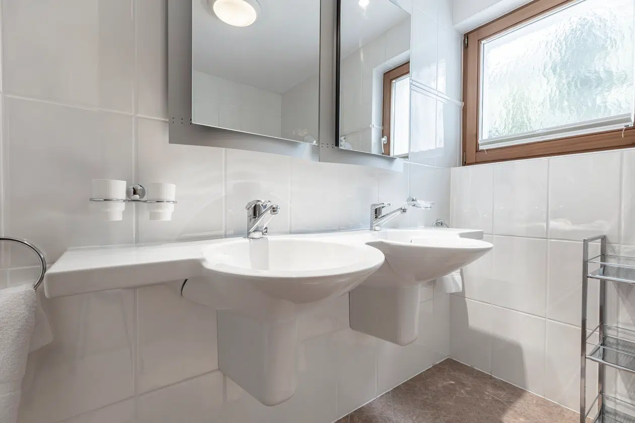 Weißes Badezimmer mit Doppelwaschbecken, Spiegeln und natürlichem Licht in der Ferienwohnung Samnaun.