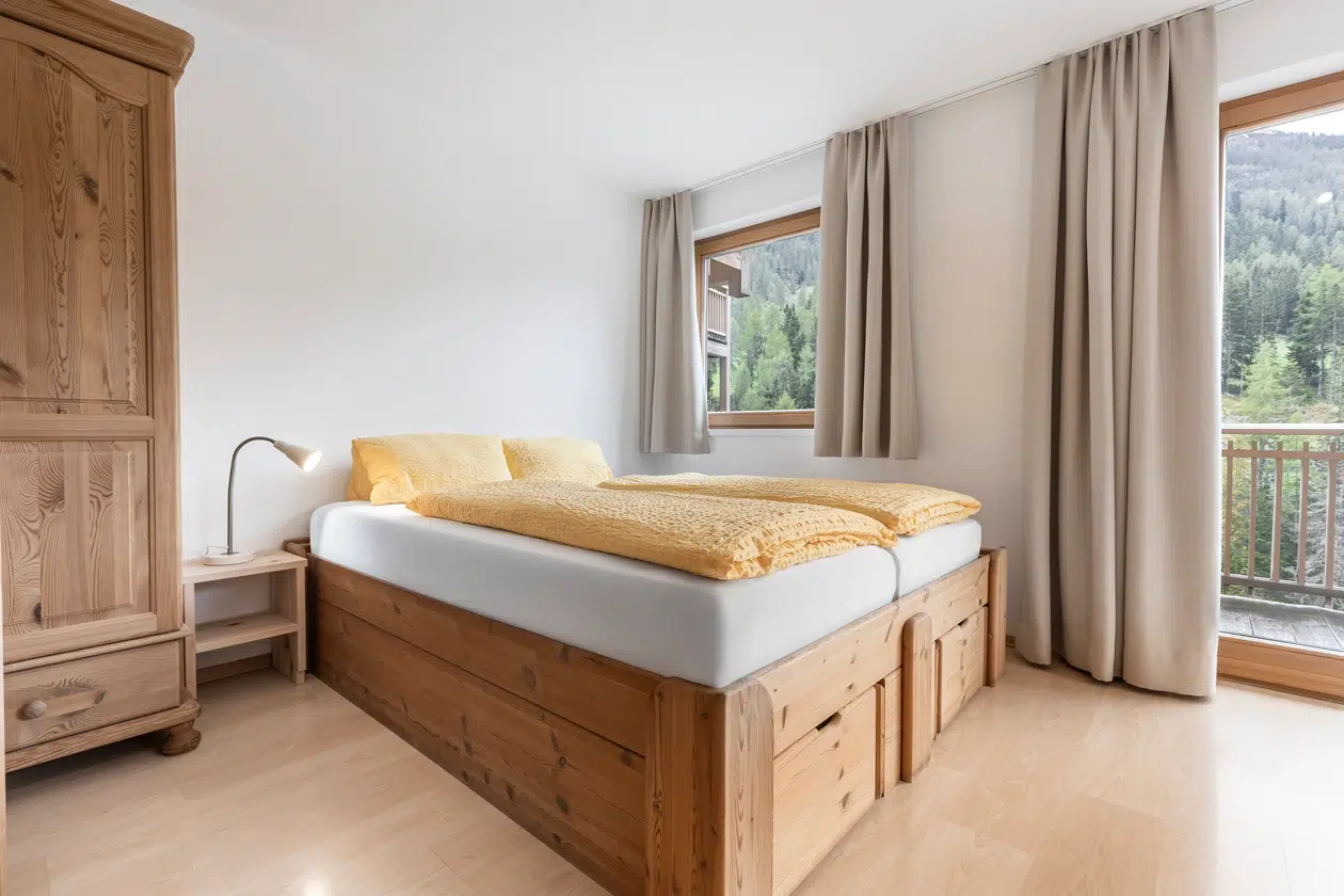 Helles Schlafzimmer in den Ferienwohnungen Samnaun mit Holzmöbeln, gelber Bettwäsche und großen Fenstern.