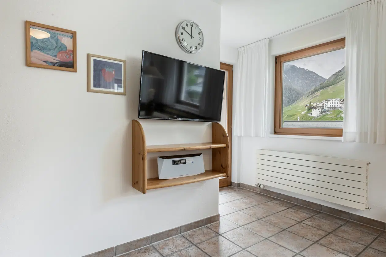 Ein an der Wand montierter Fernseher mit Regal, Kunst und Fenster mit Bergblick in der Ferienwohnung Samnaun.