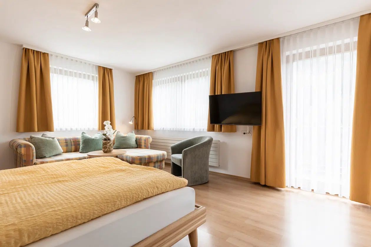 Helles Schlafzimmer in der Ferienwohnung Samnaun mit Senfvorhängen, Bett, Sofa, Stuhl und TV.