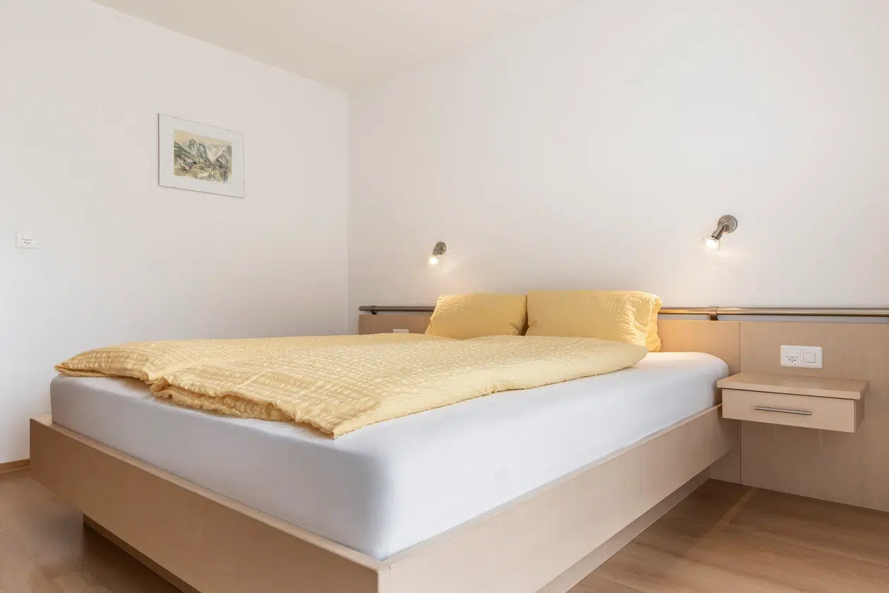 Minimalistisches Schlafzimmer in der Ferienwohnung Samnaun mit hellem Holzbett, gelbem Bettzeug und Leselampen.