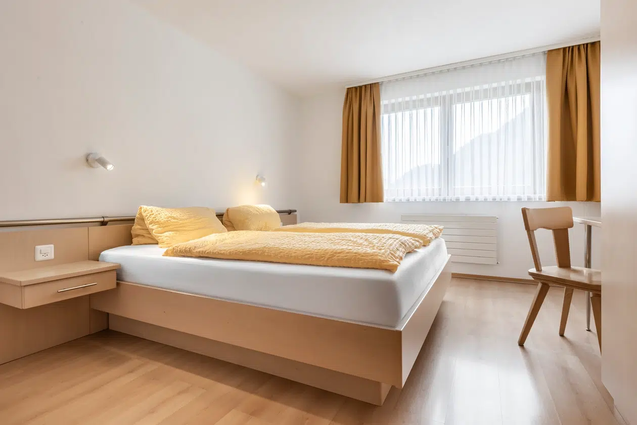 Minimalistisches Schlafzimmer in der Ferienwohnung Samnaun mit gelbem Bettzeug, Holzboden und großem Fenster.