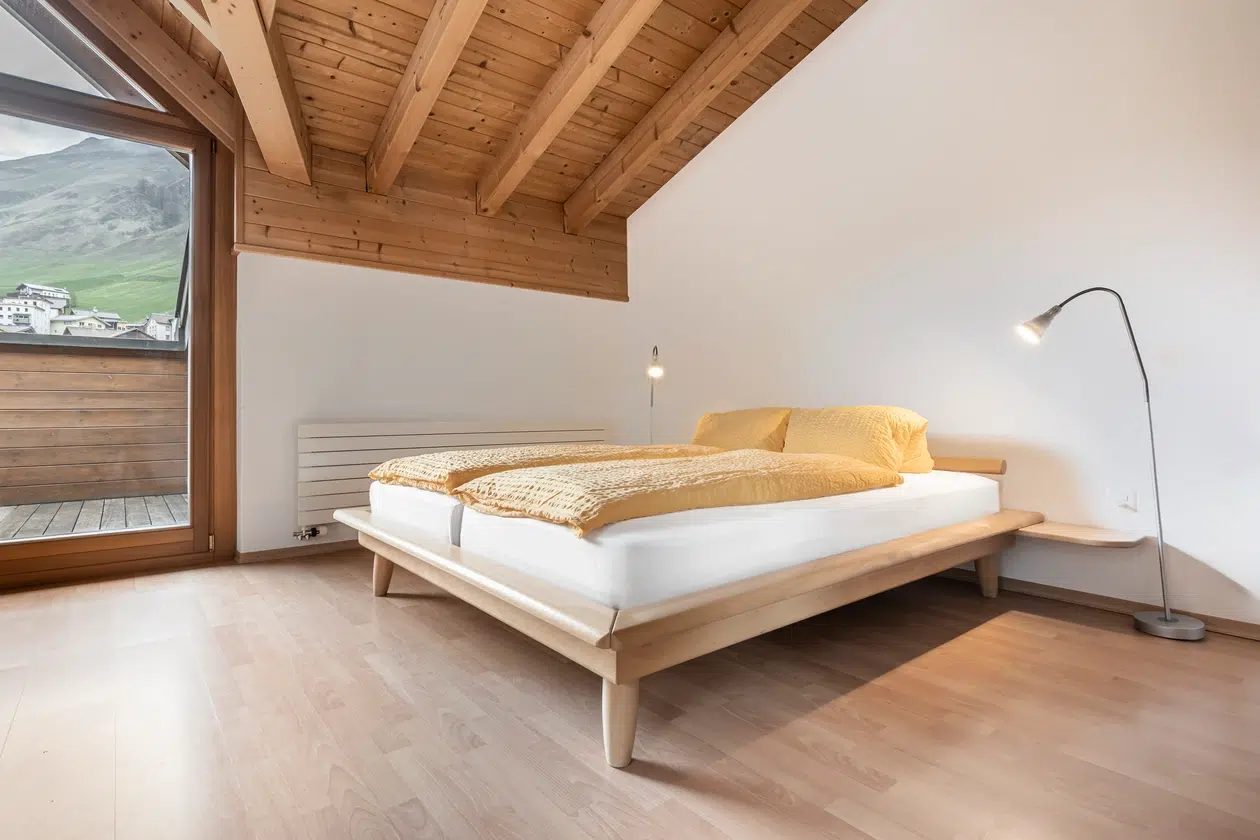 Minimalistisches Schlafzimmer in der Ferienwohnung Samnaun mit Holzdecke, gelbem Bettzeug und Bergblick.