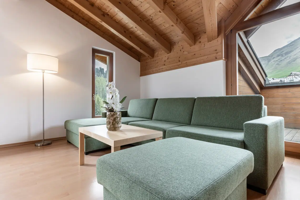 Modernes Wohnzimmer in der Ferienwohnung Samnaun mit grünem Sofa, Holzdecke und Bergblick.