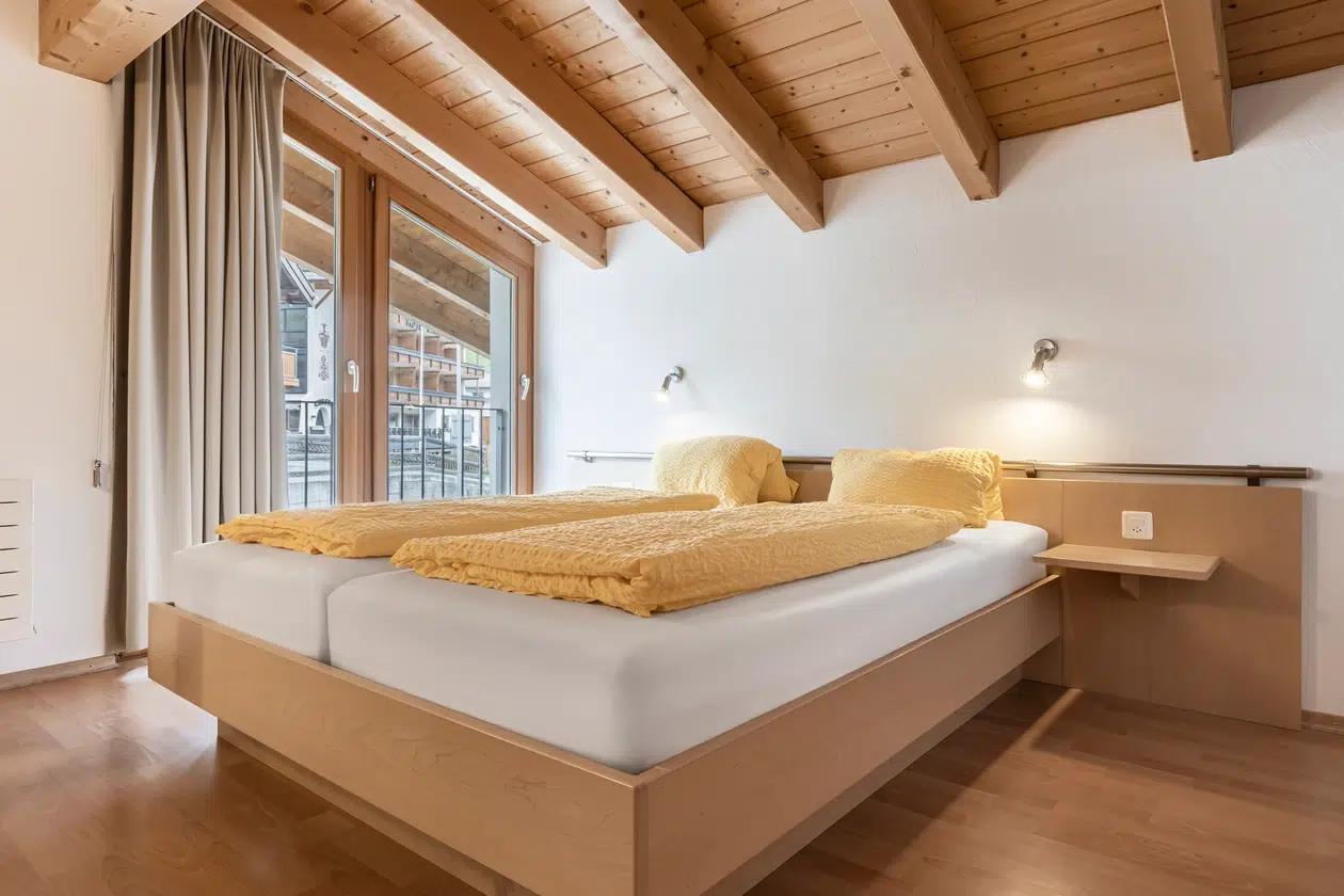 Gemütliches Schlafzimmer in der Ferienwohnung Samnaun mit zwei Betten, gelber Bettwäsche und großen Fenstern.