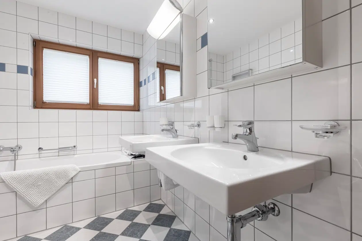 Weiß gefliestes Badezimmer in der Ferienwohnung Samnaun mit Badewanne, zwei Waschbecken und kariertem Boden.