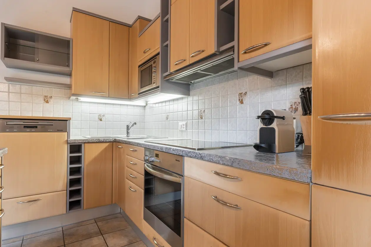 Moderne Küche mit hellen Holzschränken, Herd, Spüle und Kaffeemaschine auf grauen Arbeitsplatten.