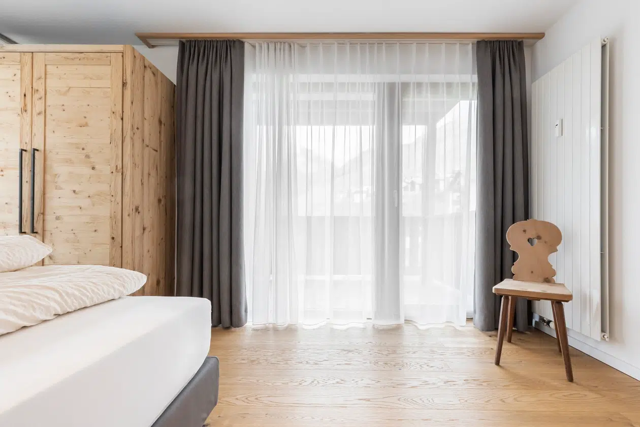 Minimalistisches Schlafzimmer mit Holzboden, Vorhängen, einem Holzstuhl und einem sichtbaren Teil eines Bettes.