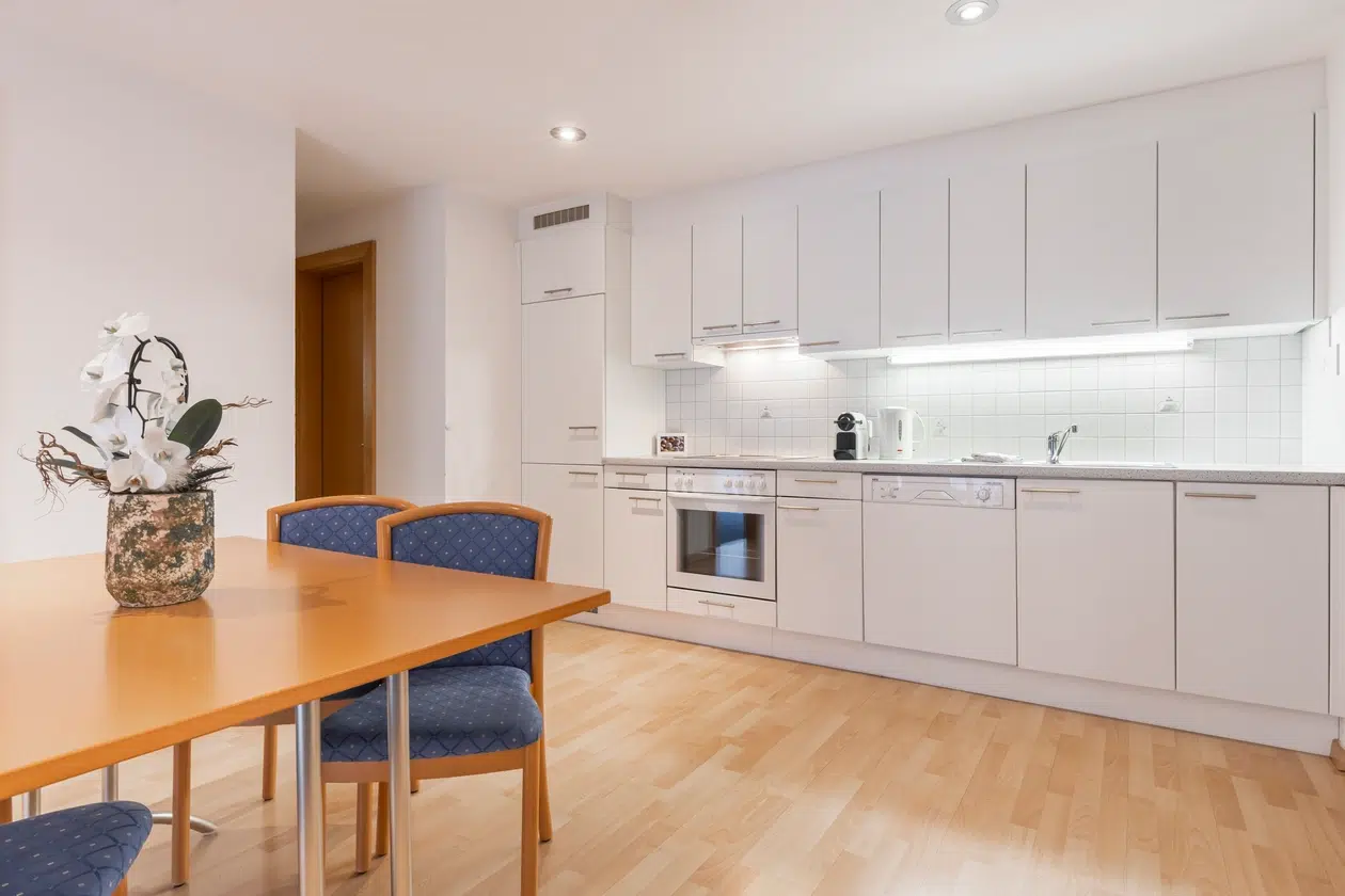 Moderne weiße Küche im Apartment Samnaun mit Holzboden, blauen Stühlen und einer Blumenvase.