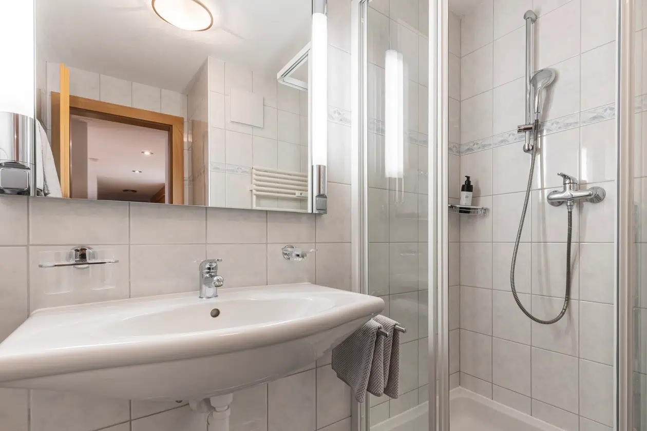 Weiß gefliestes Badezimmer im Apartment Samnaun mit Waschbecken, Spiegel, Handtuch und Glasdusche.