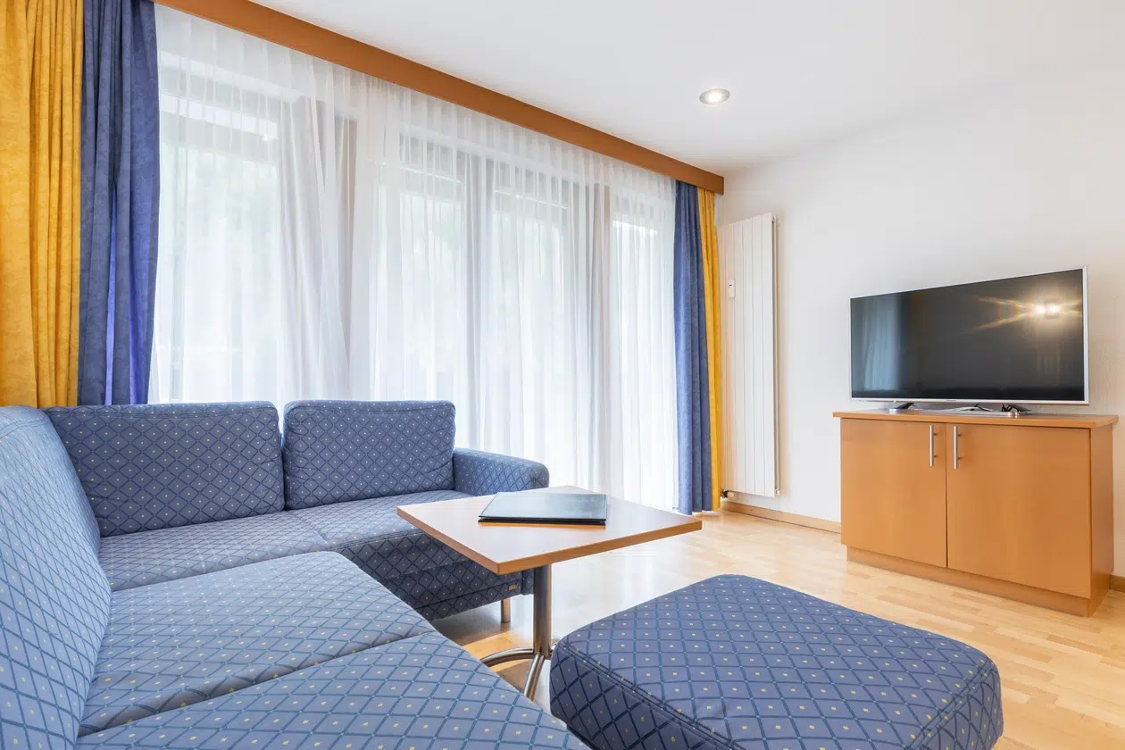 Helles Wohnzimmer im Apartment Samnaun mit blauer Couch, TV-Ständer und großen Fenstern.