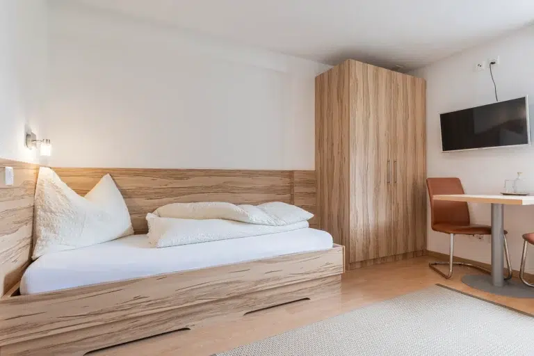 Minimalistisches Zimmer Samnaun mit Einzelbett, Holzkleiderschrank, Wand-TV und kleinem Esstisch.