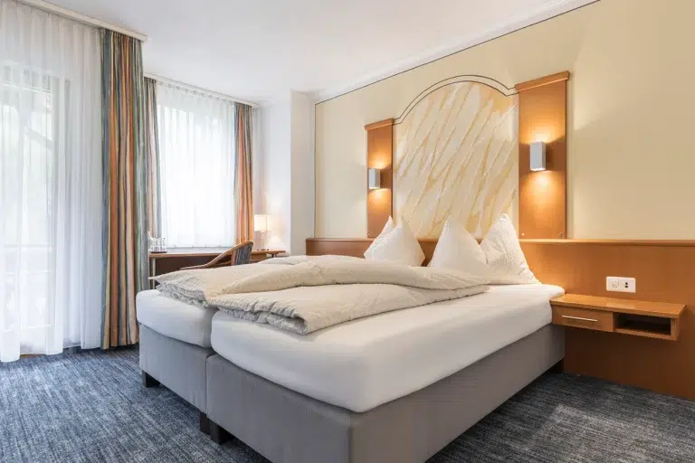 Ein modernes Doppelzimmer Samnaun mit einem Doppelbett, Schreibtisch und großen Fenstern mit Vorhängen.