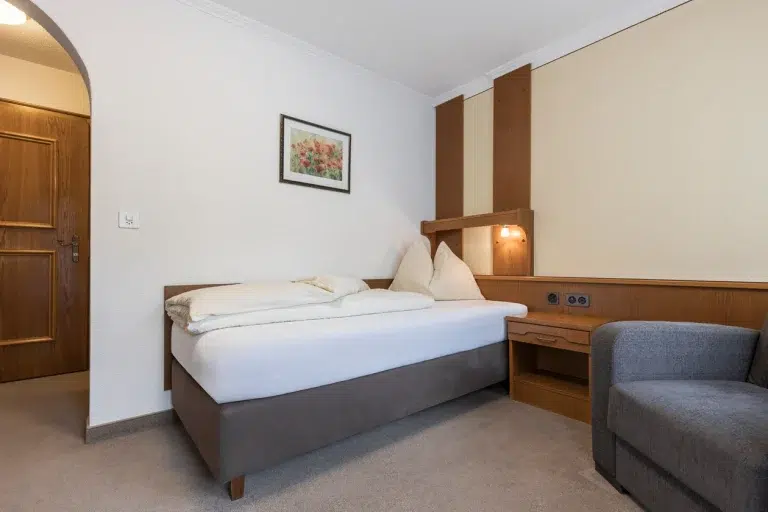 Ein aufgeräumtes Zimmer Samnaun mit einem Einzelbett, Nachttisch, Kunstwerken und einem grauen Sessel.
