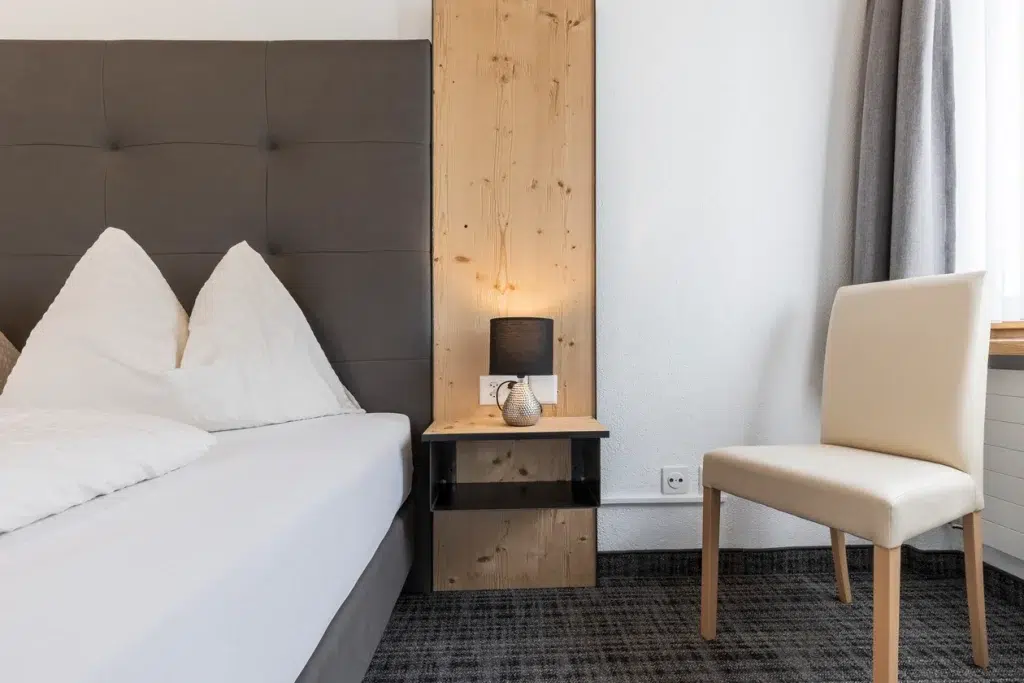 Minimalistisches Schlafzimmer im Ferienhaus Samnaun mit Bett, Nachttisch, Lampe und beigem Stuhl am Fenster.