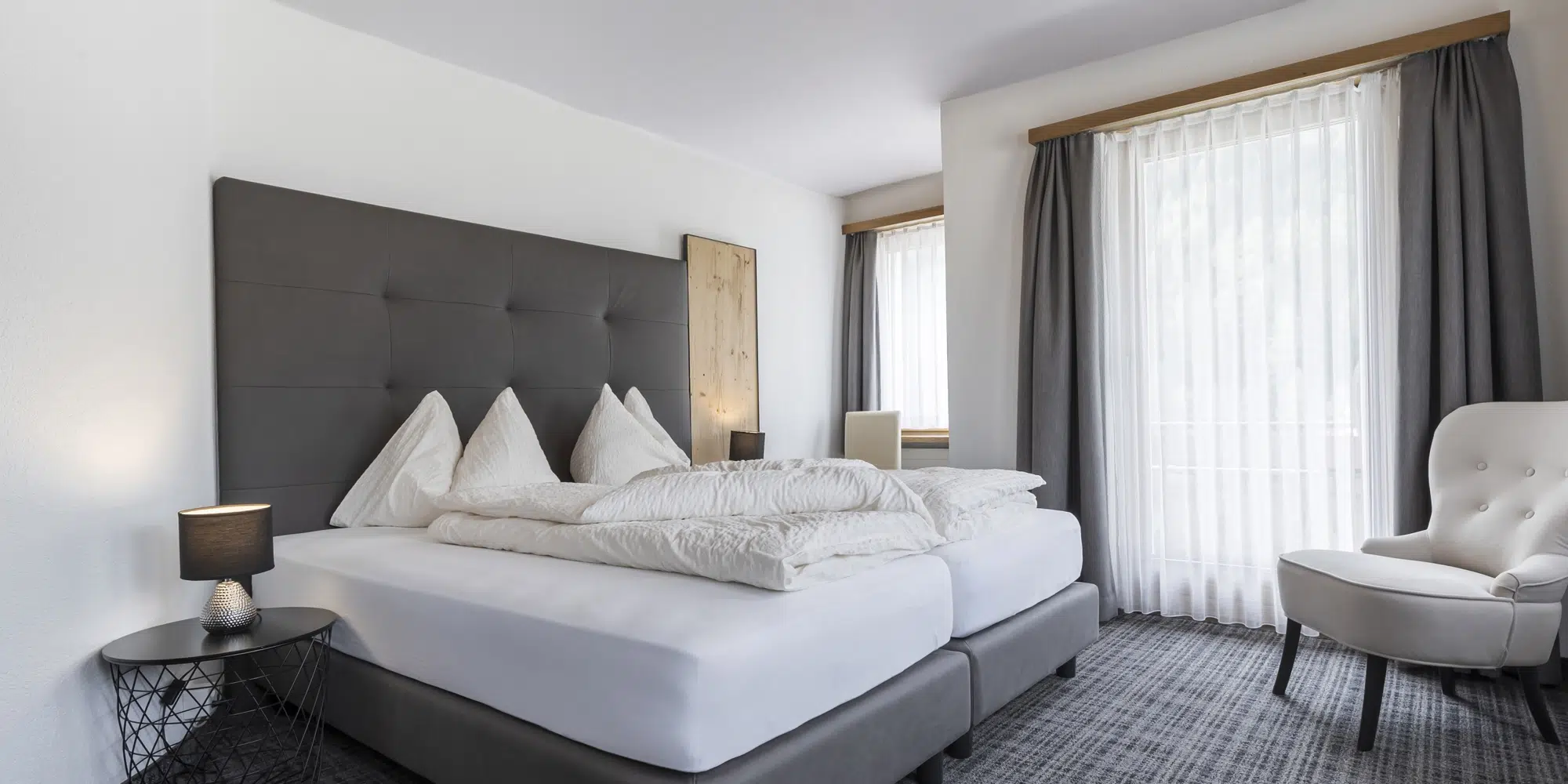 Modernes Schlafzimmer im Ferienhaus Samnaun mit großem Bett, Sessel und transparenten Fenstervorhängen.