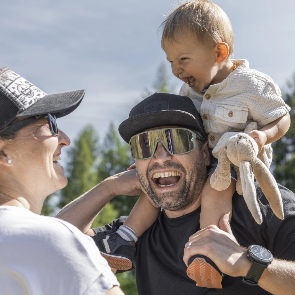 Mann mit Sonnenbrille trägt ein lächelndes Kind auf seinen Schultern in der Nähe von unterkunft Samnaun im Freien.