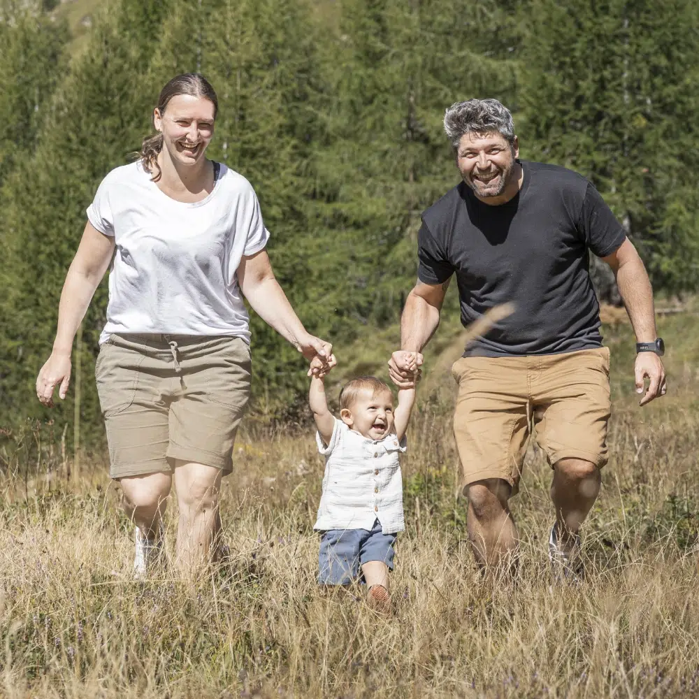 Lächelnde Erwachsene, die ein Kind an der Hand halten, während sie durch ein grasbewachsenes Feld mit Bäumen dahinter gehen.