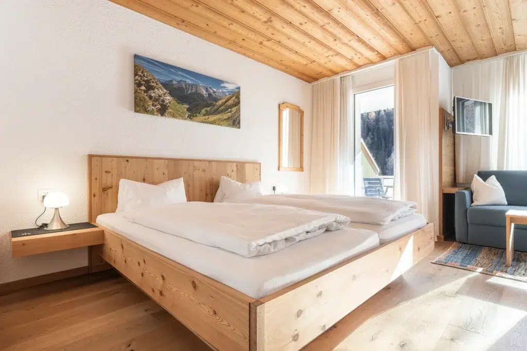 Helles Zimmer im Apartment Samnaun mit Holzbett, weißem Bettzeug und Sonnenlicht durch große Fenster.