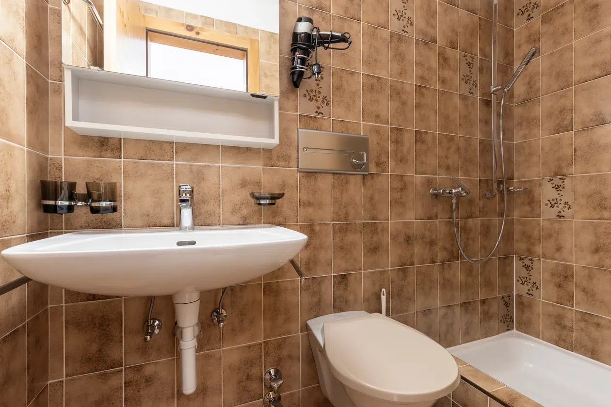 Kleines Badezimmer im Apartment Samnaun mit braun gefliesten Wänden, Waschbecken, Toilette, Spiegel und Dusche.