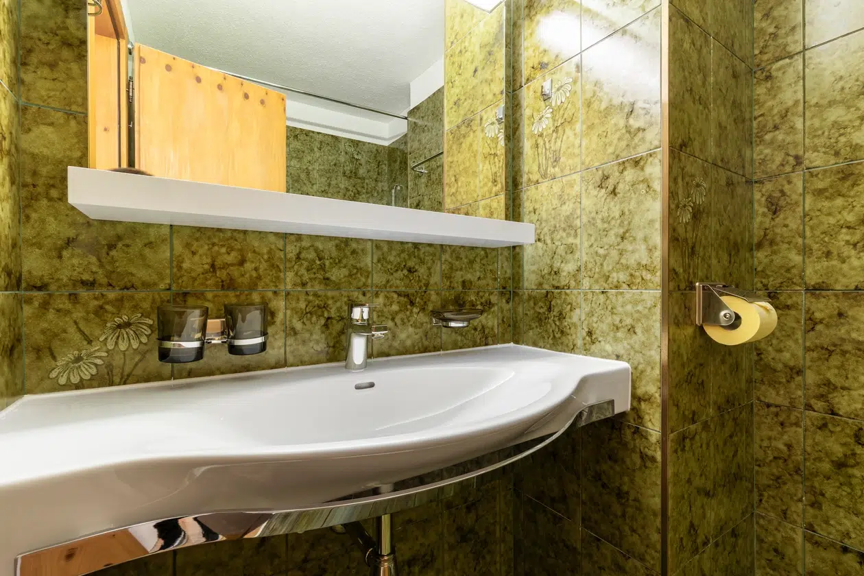 Weißes Waschbecken mit verchromtem Wasserhahn im Badezimmer von Apartment Samnaun mit grünlichen Fliesen und einem Spiegel.