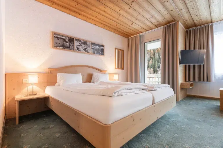 Helles Apartment Samnaun Zimmer mit großem Bett, Holzdecke, Lampen, TV und großen Fenstern.
