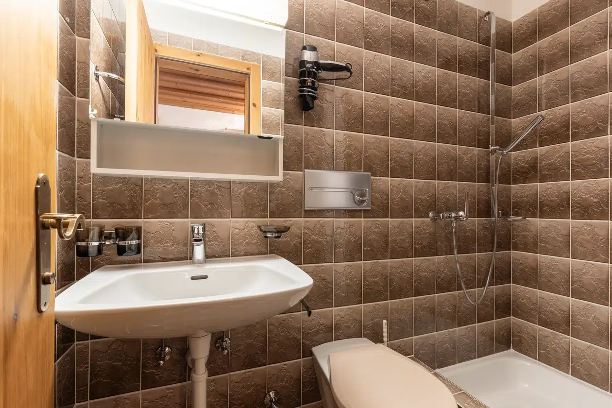 Kleines Badezimmer im Apartment Samnaun mit braunen Fliesen, weißem Waschbecken, Toilette, Spiegel und Dusche.