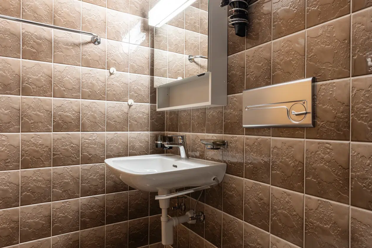 Kleines Badezimmer im Apartment Samnaun mit braun gemusterten Fliesen, weißem Waschbecken und Spiegel.