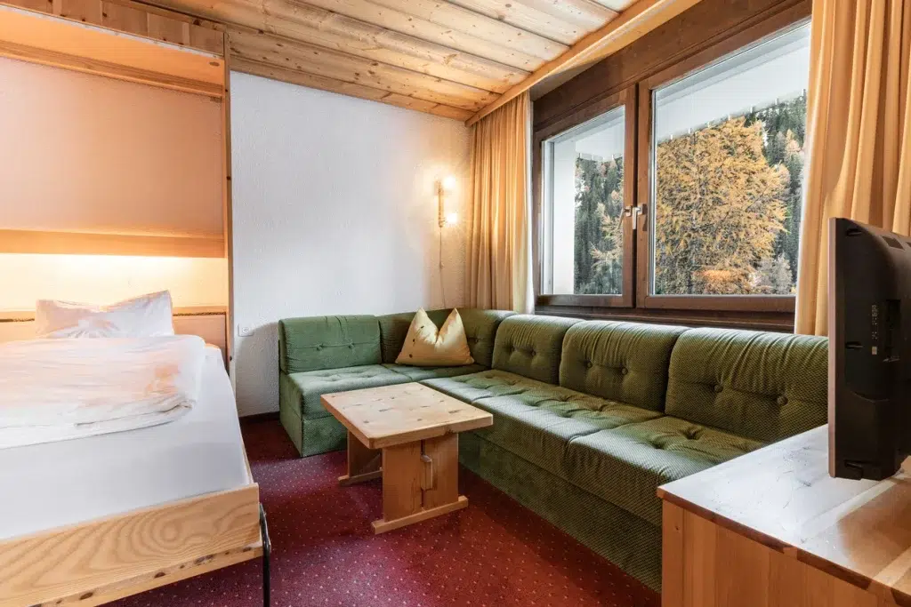 Gemütliches Zimmer im Apartment Samnaun mit grünem Sofa, Holzakzenten, Bett und Fenster mit Blick auf Bäume.