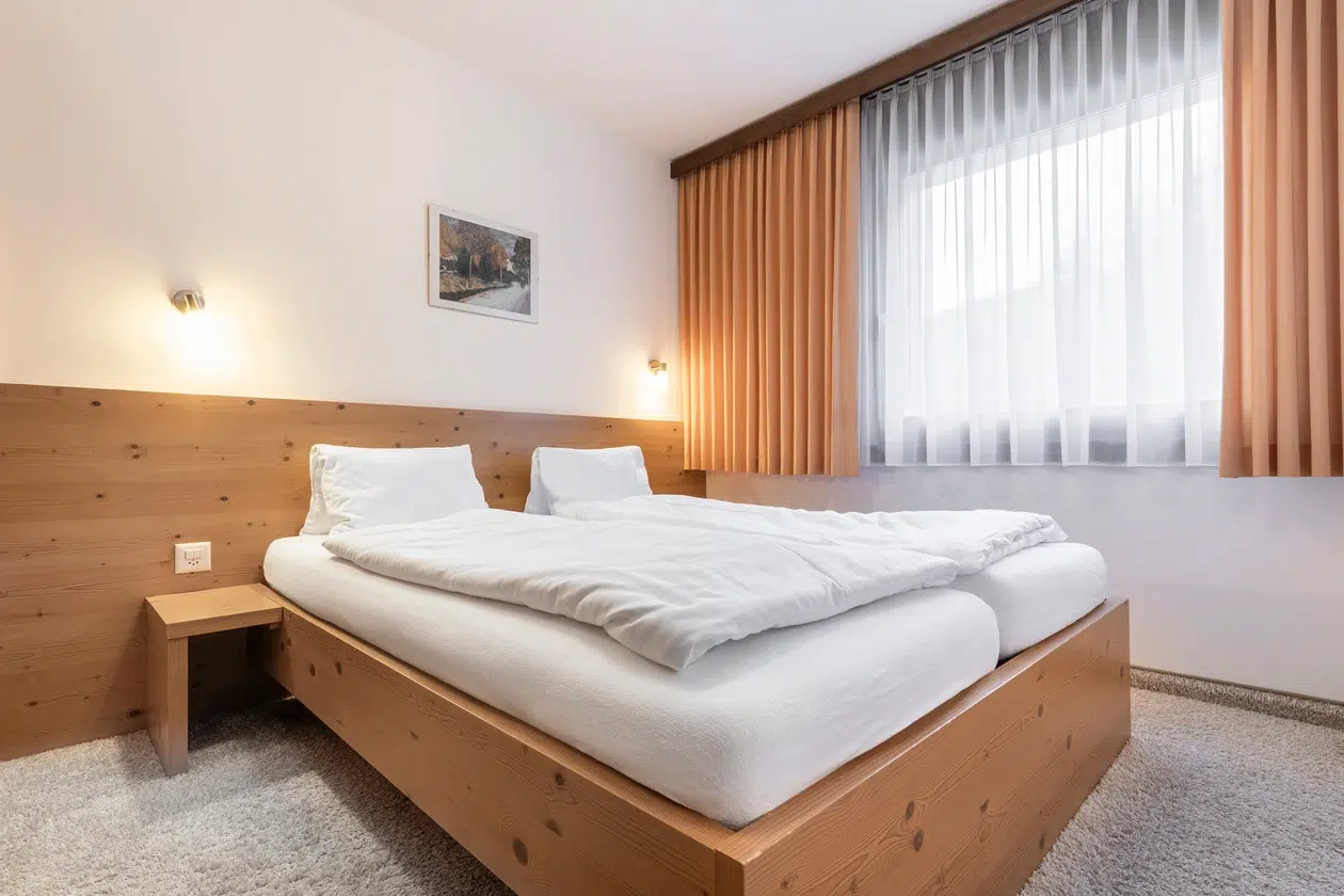 Minimalistisches Schlafzimmer im Apartment Samnaun mit einem Holzbett, weißer Bettwäsche und beigen Vorhängen.