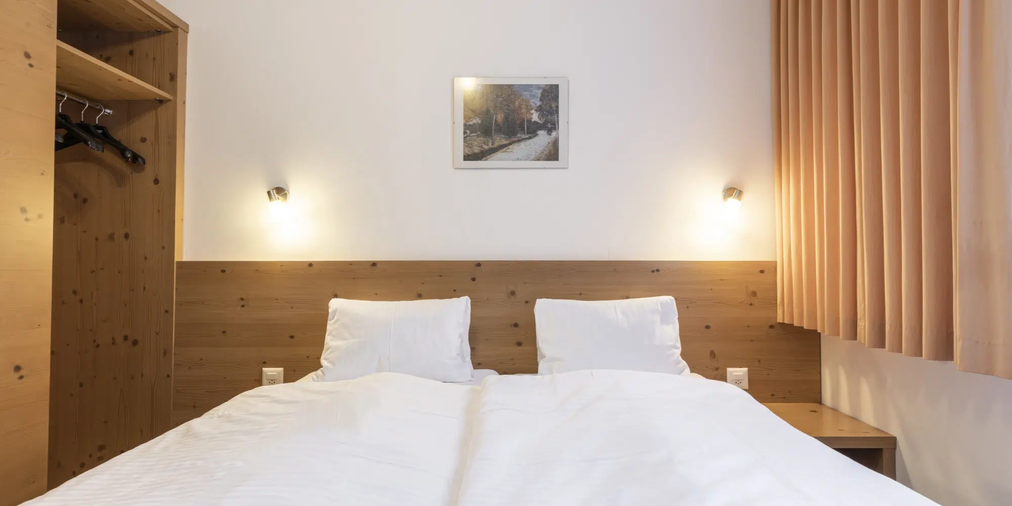 Gepflegtes Doppelbett mit weißer Bettwäsche im Ferienhaus Samnaun, Holzkopfteil, Wandlampen.