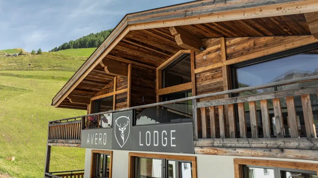 Ferienhaus Samnaun aus Holz mit Hirsch-Logo inmitten von grünen Hügeln und blauem Himmel.