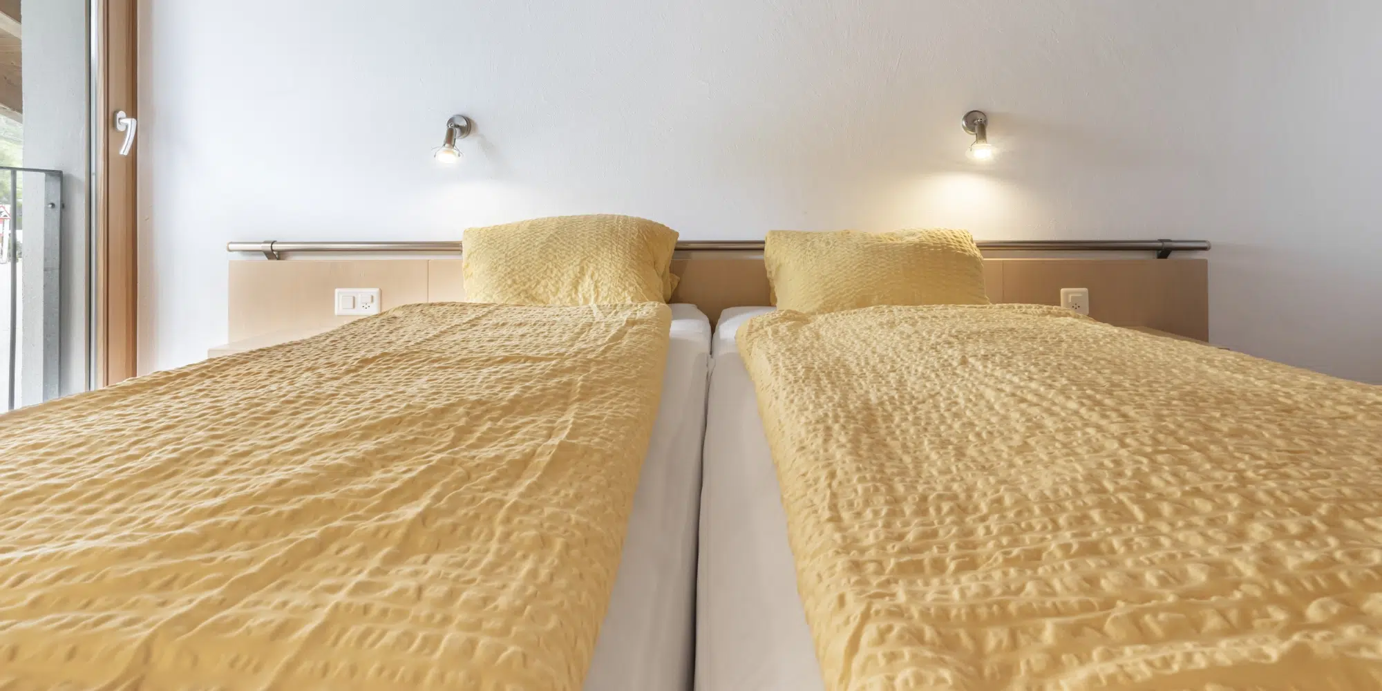 Zwei Einzelbetten mit gelber Bettwäsche in den Ferienwohnungen Samnaun, jeweils mit Wandleselampen.