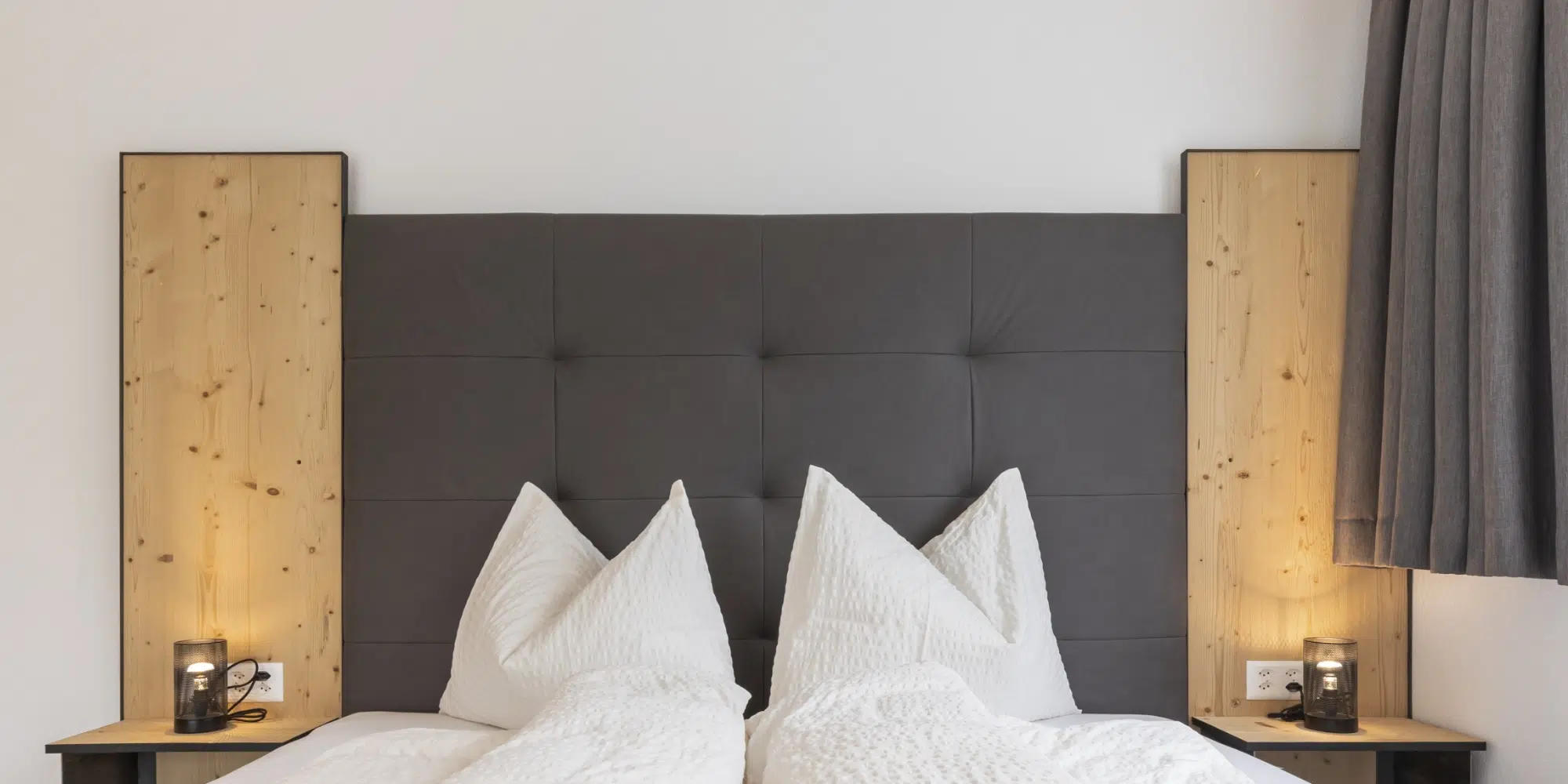 Modernes Bett im ferienhaus Samnaun mit weißen Kissen, grauem Kopfteil und Nachttischlampen.