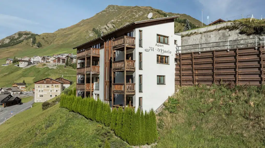Modernes Alpen-Ferienhaus Samnaun mit Holzbalkonen an einem grasbewachsenen Hang, Berge im Hintergrund.