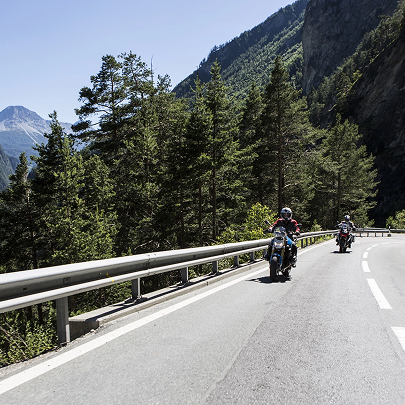 Zwei Motorradfahrer fahren auf einer kurvenreichen Bergstraße bei Samnaun, umgeben von Bäumen und Hügeln.