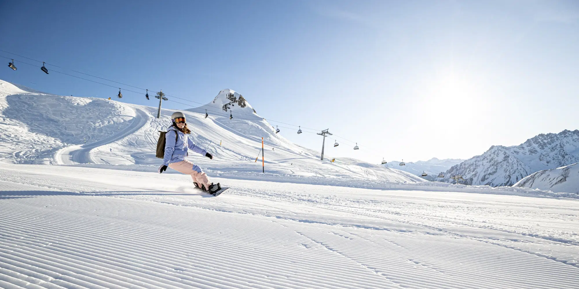 Eine Person beim Snowboarden auf einer präparierten Schneepiste in Samnaun, mit Skiliften und Bergen dahinter.