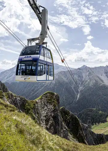 A cable car travels over a mountainous landscape with grassy slopes and distant peaks.