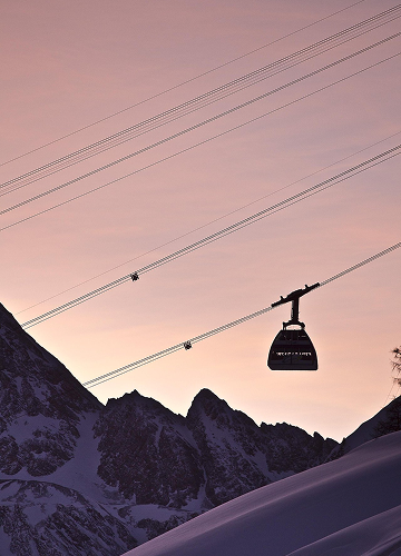 Seilbahn vor rosafarbenem Himmel, mit schneebedeckten Berggipfeln im Hintergrund.