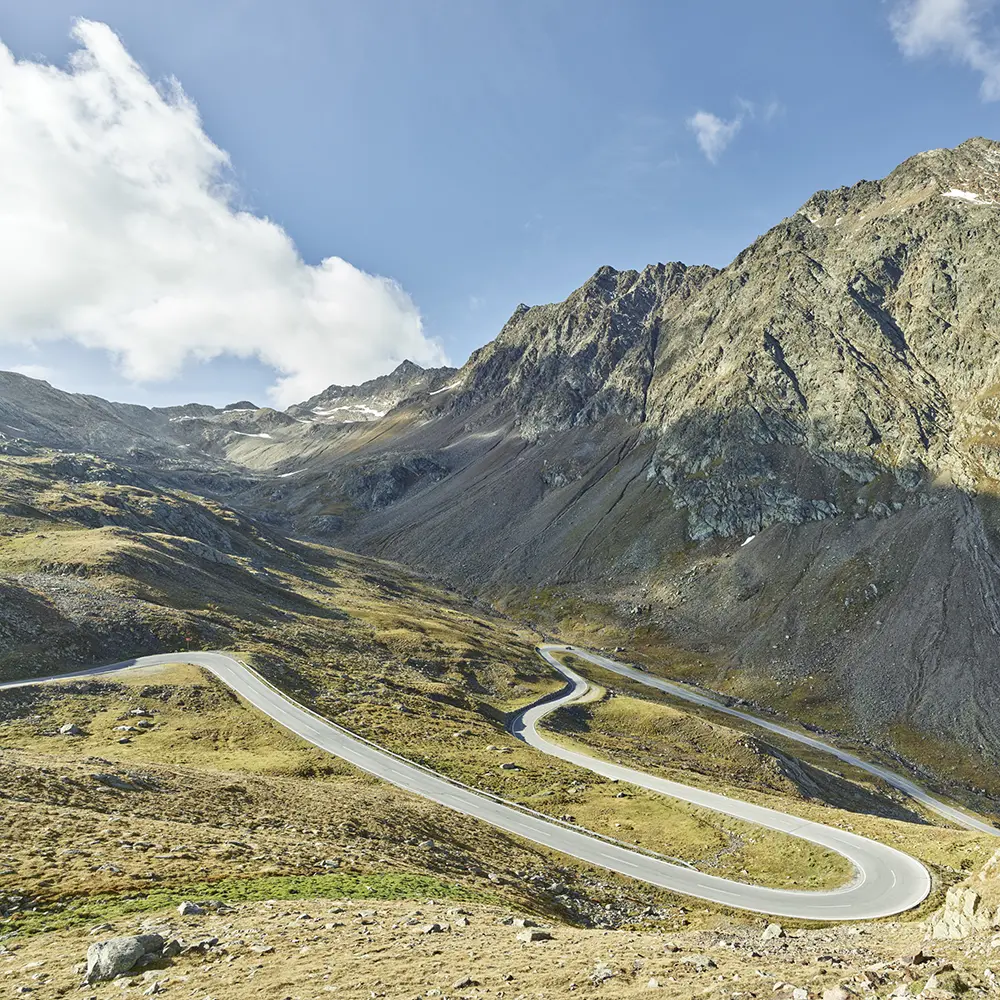 Eine kurvenreiche Bergstraße in Samnaun schlängelt sich durch felsige, grasbewachsene Hügel unter blauem Himmel.