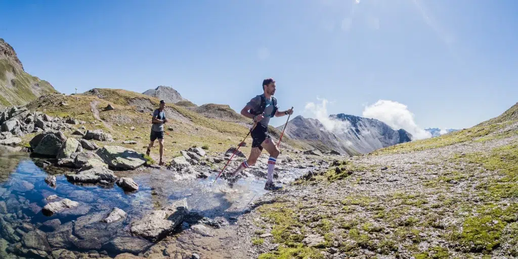 Zwei Trailrunner mit Stöcken überqueren einen felsigen Bach in den sonnenbeschienenen Samnauner Bergen.