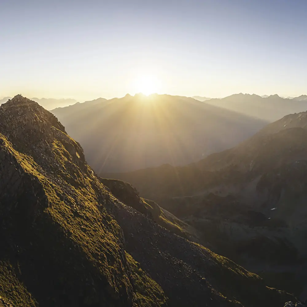 Die Sonne geht über den fernen Samnauner Berggipfeln mit einem felsigen Abhang im Vordergrund auf.